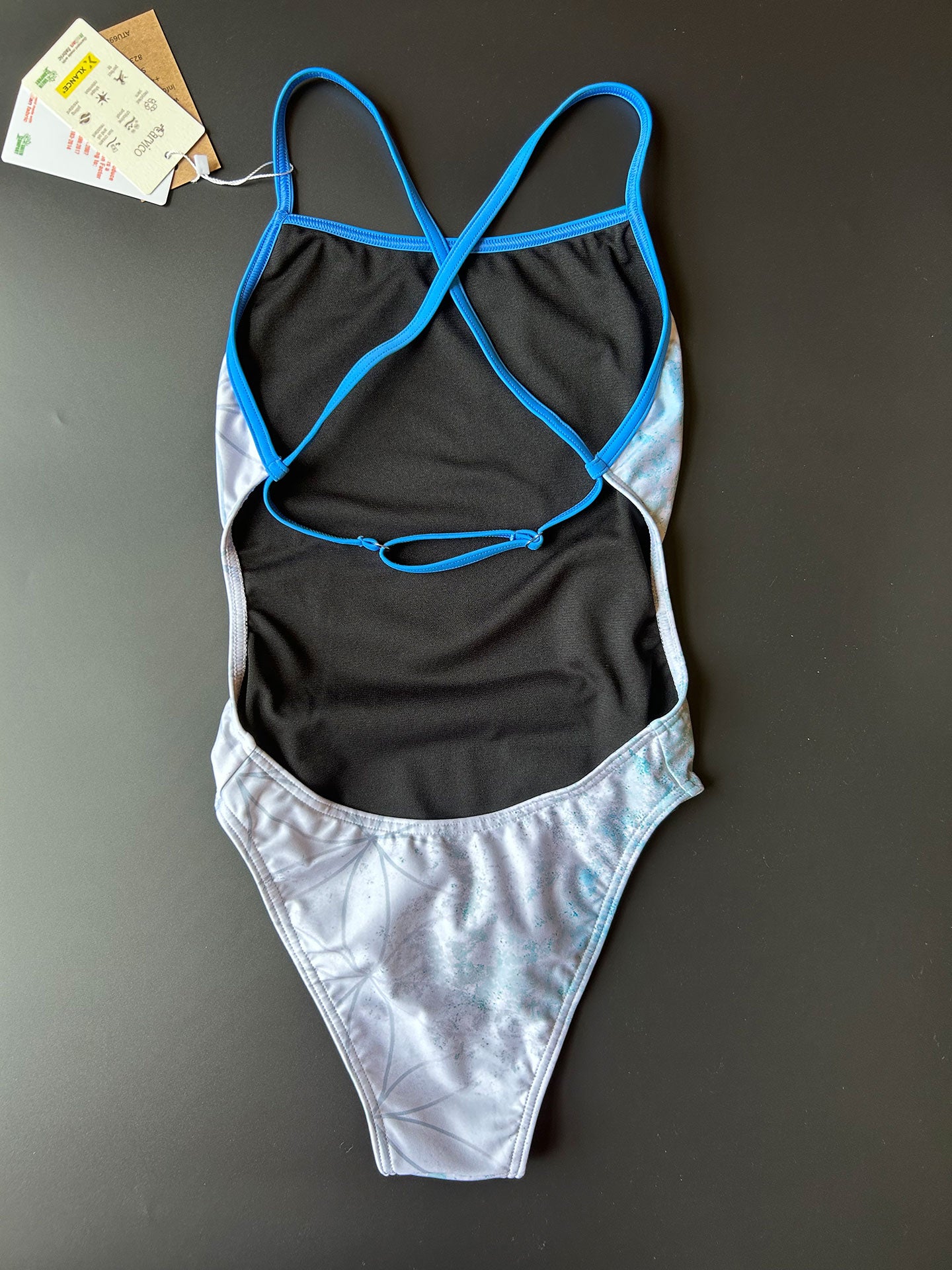 FLOWER OF LIFE - One-Piece Swimsuit lying on a black surface and shown from the back with its open back and thin blue adjustable straps.