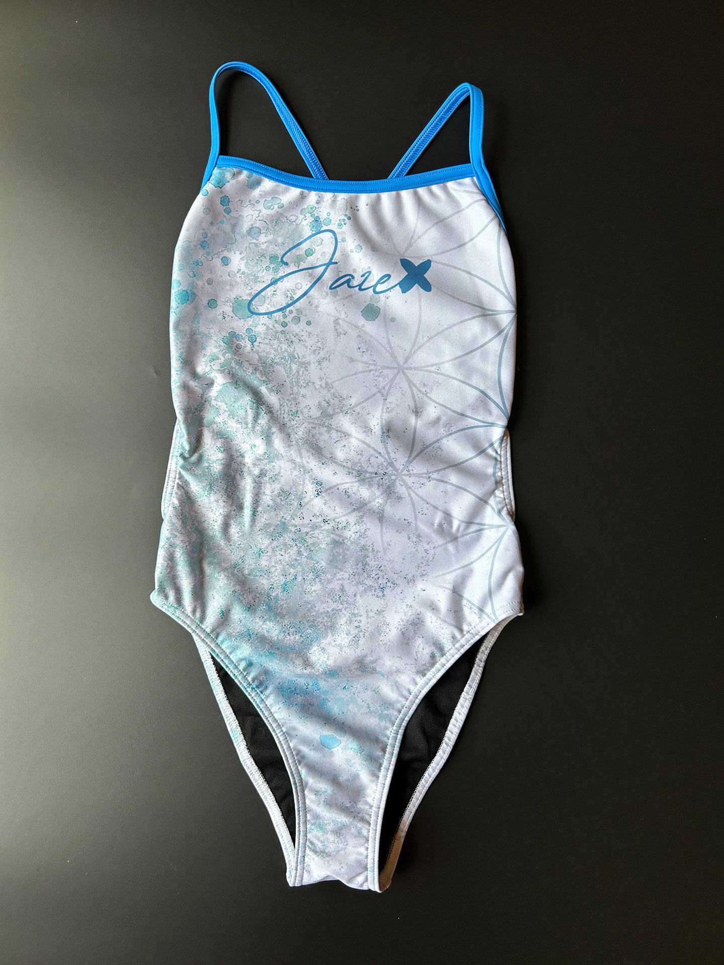 FLOWER OF LIFE - One-Piece Swimsuit lying on a black surface showing the high cut leg and the blue straps.