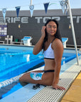 Female model wearing the Flower of life – Bikini top while sitting by the pool, a swim cap on her lap.
