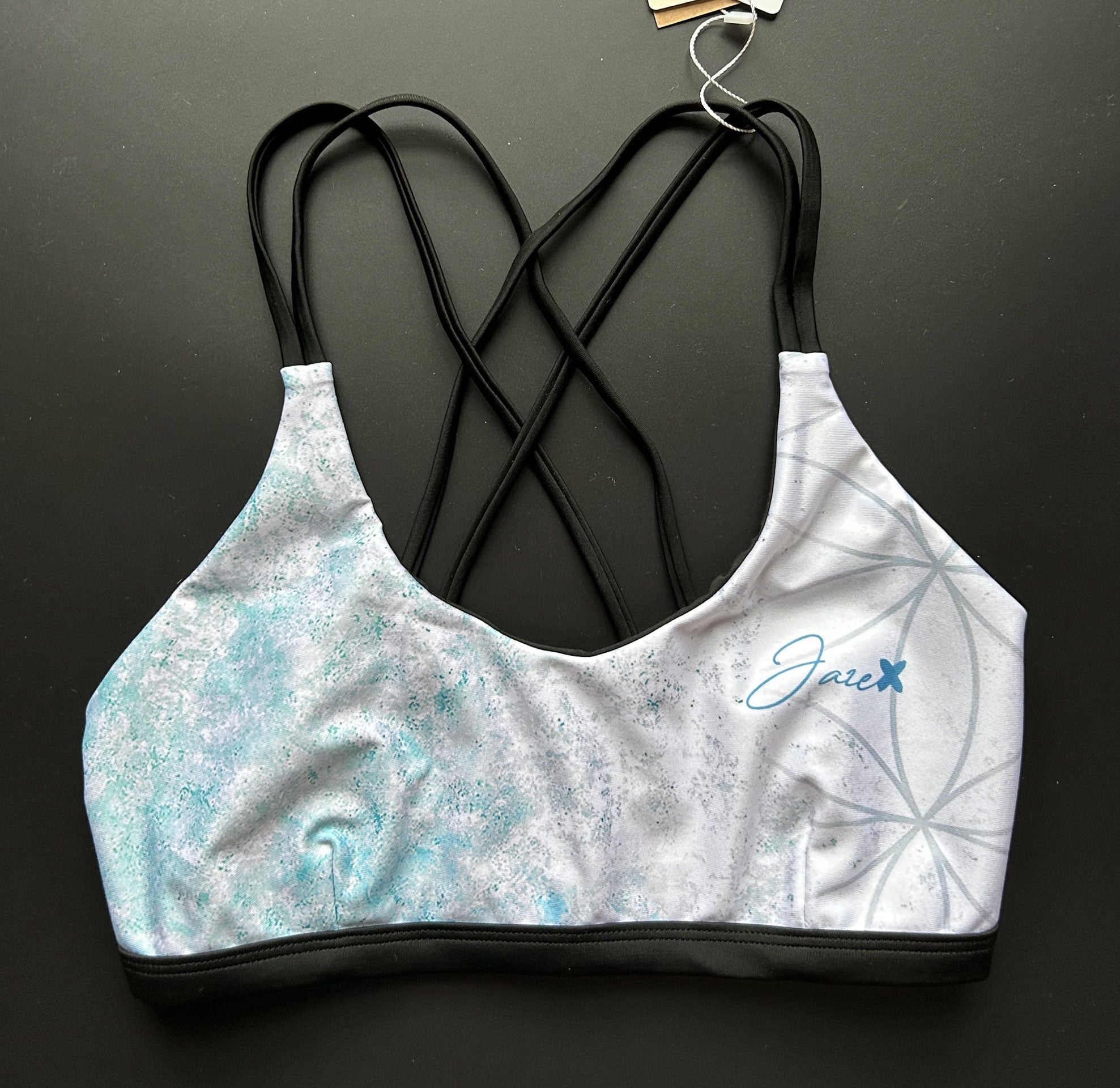 Flower of life – Bikini top shown from the front lying on a black surface, focussing on the light blue patterns and thin, black stripes.