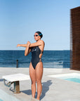Female model wearing the Hailey One-Piece Swimsuit, warming up by the pool in a side view, emphasizing the swimsuit's sporty fit.