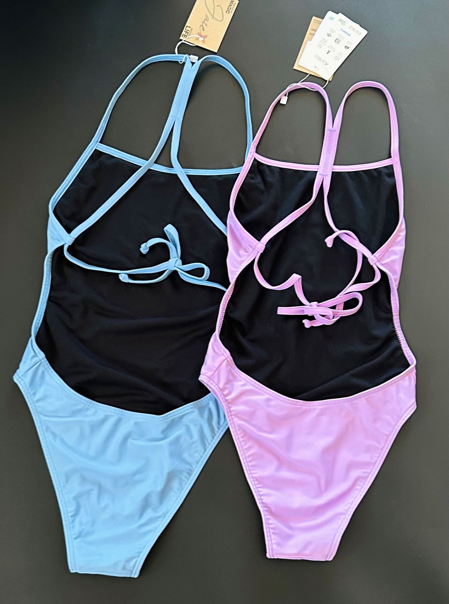 Our Lavender One-Piece-Swimsuit lying on a black surface and shown from the back, together with our into the blue one piece swimsuit.
