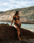 Our female Model shown from the front with our Midnight – Bikini top in dark gray, sitting on a big stone at the beach, looking to the sea.