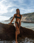 Our female Model shown from the front with our Midnight – Bikini top in dark gray, sitting on a big stone at the beach smiling confidently in the camera.
