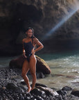 Our female Model standing on the beach and shown from the front, wearing the midnight – one piece swimsuit in dark gray. It has thin straps, a high leg cut and the white Jaze-Logo at the front left hip.