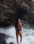 Our Model standing on the beach and shown from the back, wearing our midnight – one piece swimsuit with thin straps to tie in the back for your ultimate fit.
