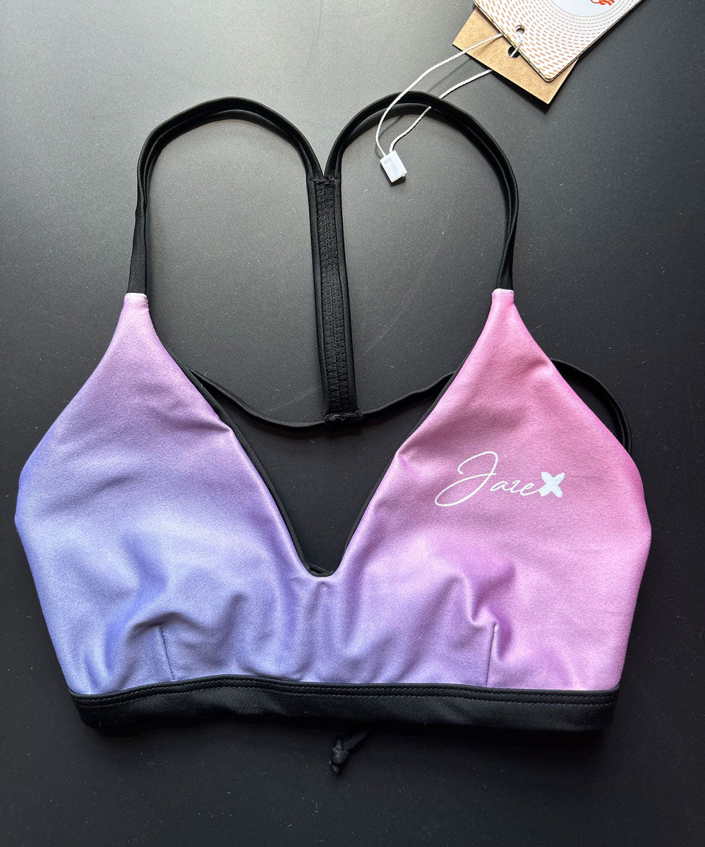 Our RAINBOW Bikini Top shown from the front and lying on a black table for more details. It is a a colorful Swimsuit with light rose and violet colors and thin, black straps.