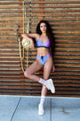 Our female model wearing our rainbow bikini bottom combined with the rainbow bikini top, leaning on a wood wall, looking confidently into the camera.