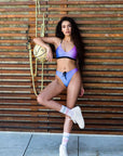 Our female model wearing our rainbow bikini bottom combined with the rainbow bikini top, leaning on a wood wall, looking confidently into the camera.