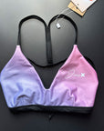 Our RAINBOW Bikini Top shown from the front and lying on a black table for more details. It is a a colorful Swimsuit with light rose and violet colors and thin, black straps.