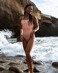 Female model wearing the Sunrise Swim Onesie at the sea, smiling and pointing to the left, showcasing the swimsuit's vibrant design and flattering fit.
