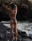 Back view of a female model in the Universe Swim Onesie at the sea, touching her hair and highlighting the swimsuit's open-back design.