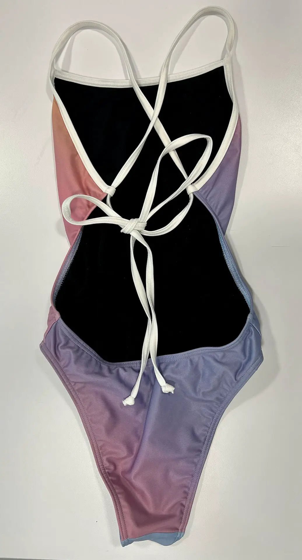 Our RAINBOW - One-Piece Swimsuit lying on a white table, where the colors and the white, thin straps can be seen in more detail. The straps in the back are tied into a stitch.