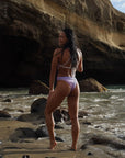 Our female Model standing on the beach and shown from the back, confidently smiling, wearing our RAINBOW - One-Piece Swimsuit with thin straps to tie in the back for your ultimate fit.