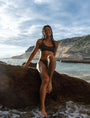 Our female Model shown from the front with our Midnight – Bikini top in dark gray, sitting on a big stone at the beach smiling confidently in the camera.