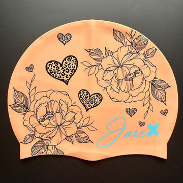 The BLOOM – swim cap has a peach base and beautiful flowers and hearts painted with fine, black lines.